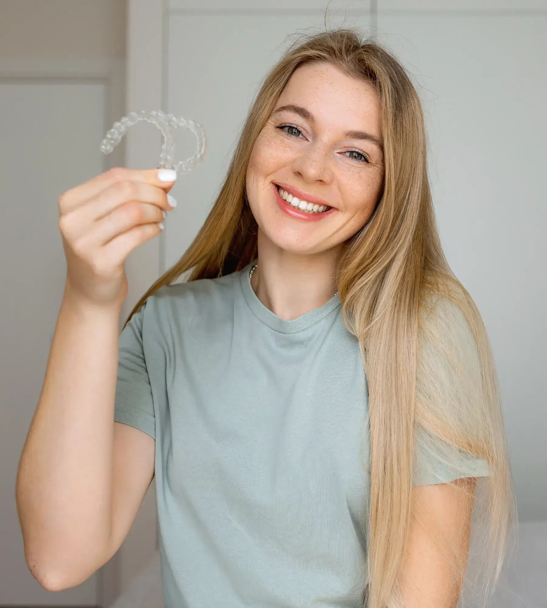 Smiling woman with long blonde hair holding clear dental aligners in front of a neutral background.