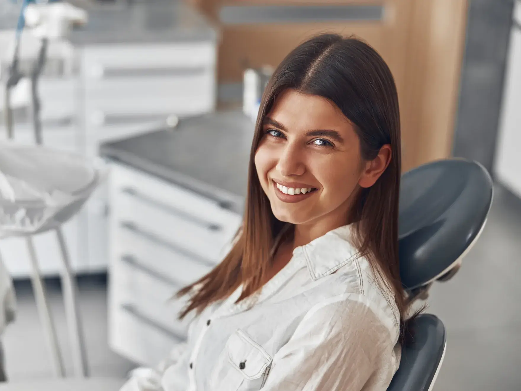 Young woman with long brown hair smiling while sitting in a dental chair in a clinic.