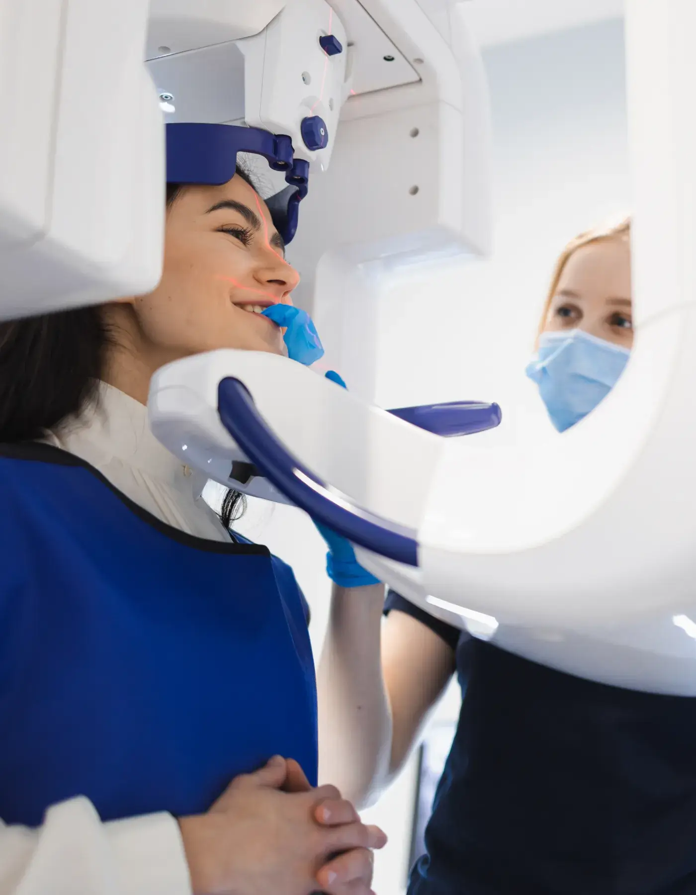 Woman wearing a blue lead apron undergoing a dental X-ray with a technician adjusting the machine while wearing a face mask and blue gloves.