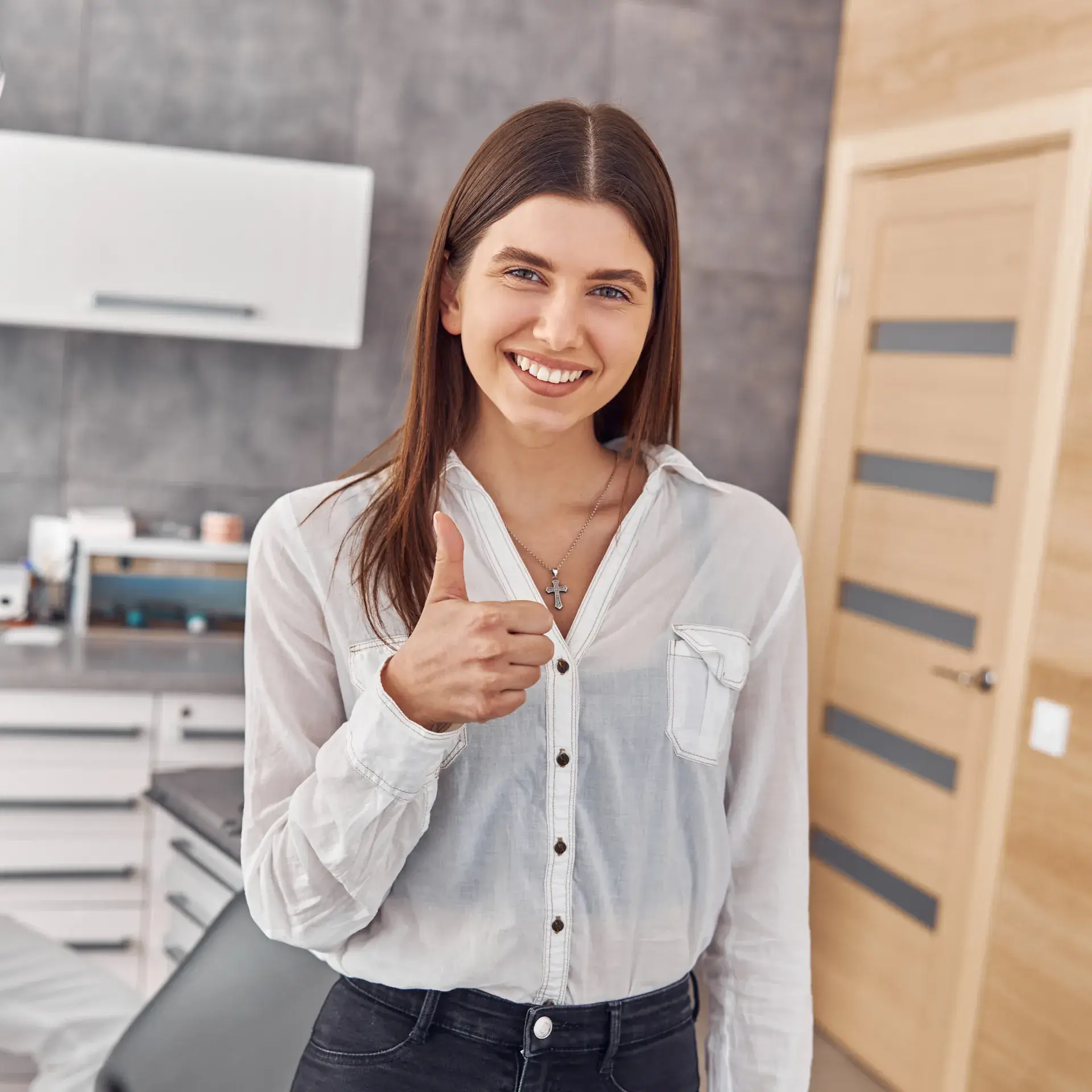 Smiling young woman wearing a white shirt giving a thumbs up in a modern room.