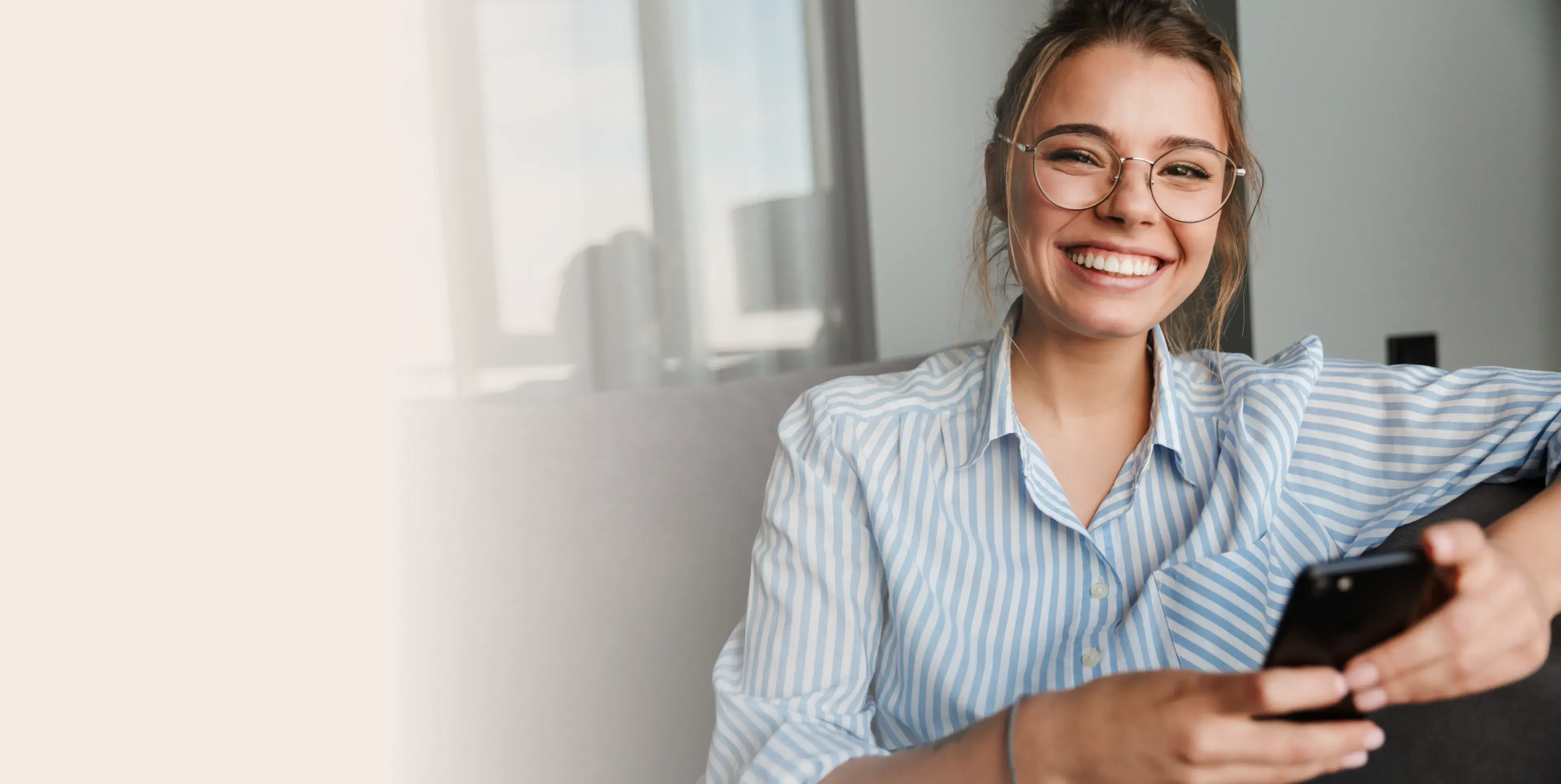 Smiling woman wearing glasses and a striped shirt holding a smartphone while sitting on a couch.