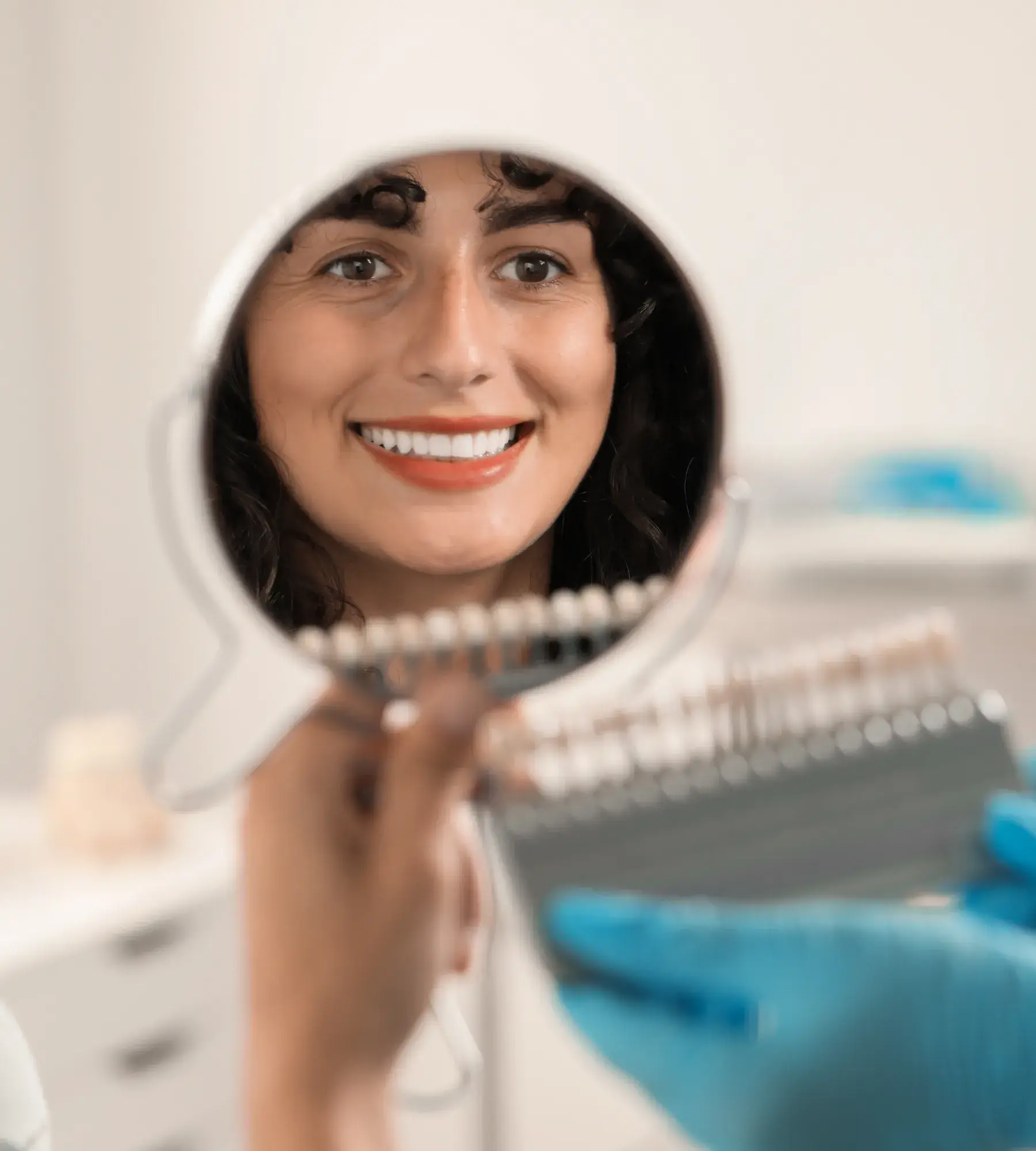 Woman smiling at her reflection in a handheld mirror while a gloved hand holds a dental shade guide next to her teeth.
