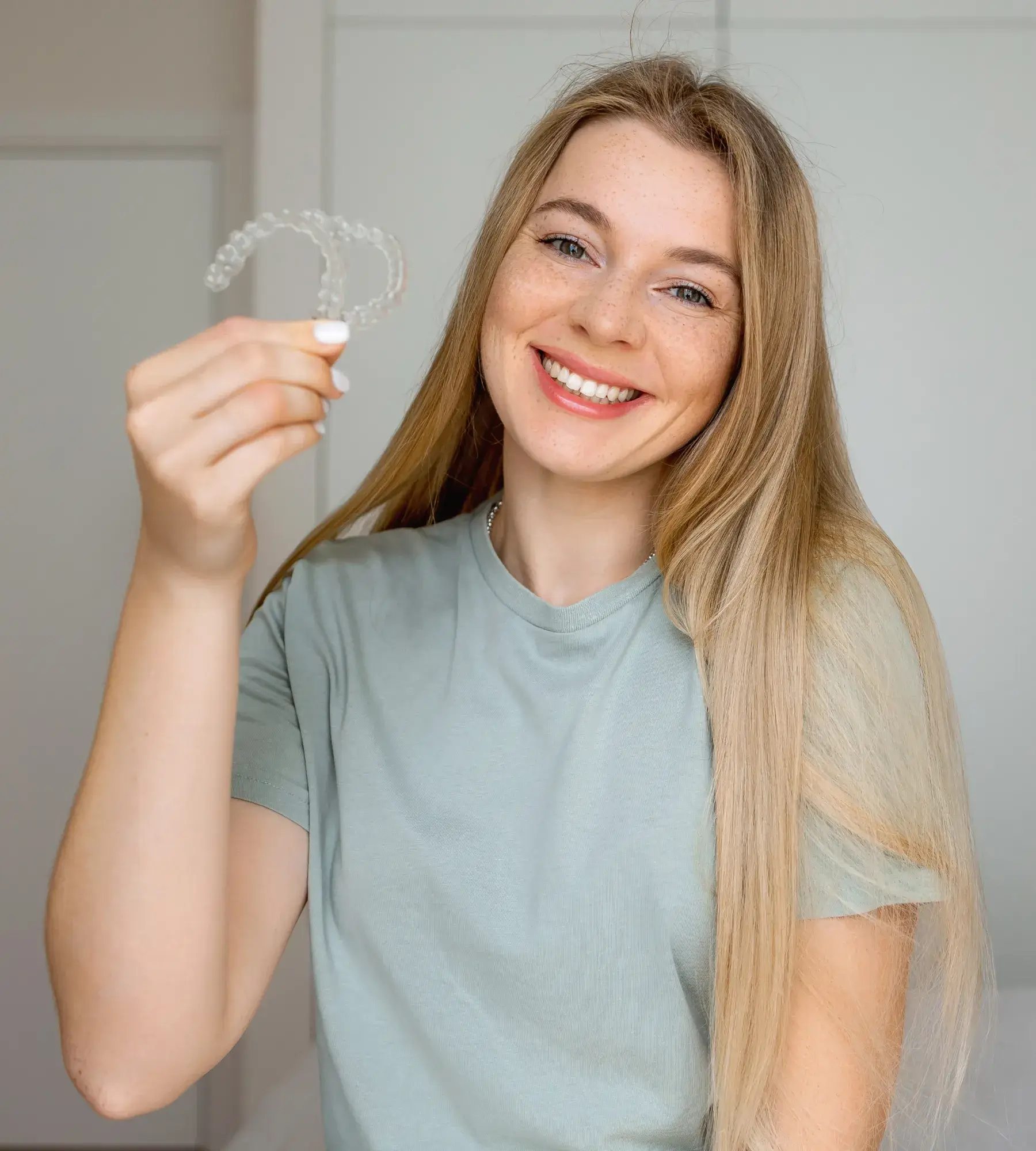 Smiling young woman with long blonde hair holding clear dental aligners.
