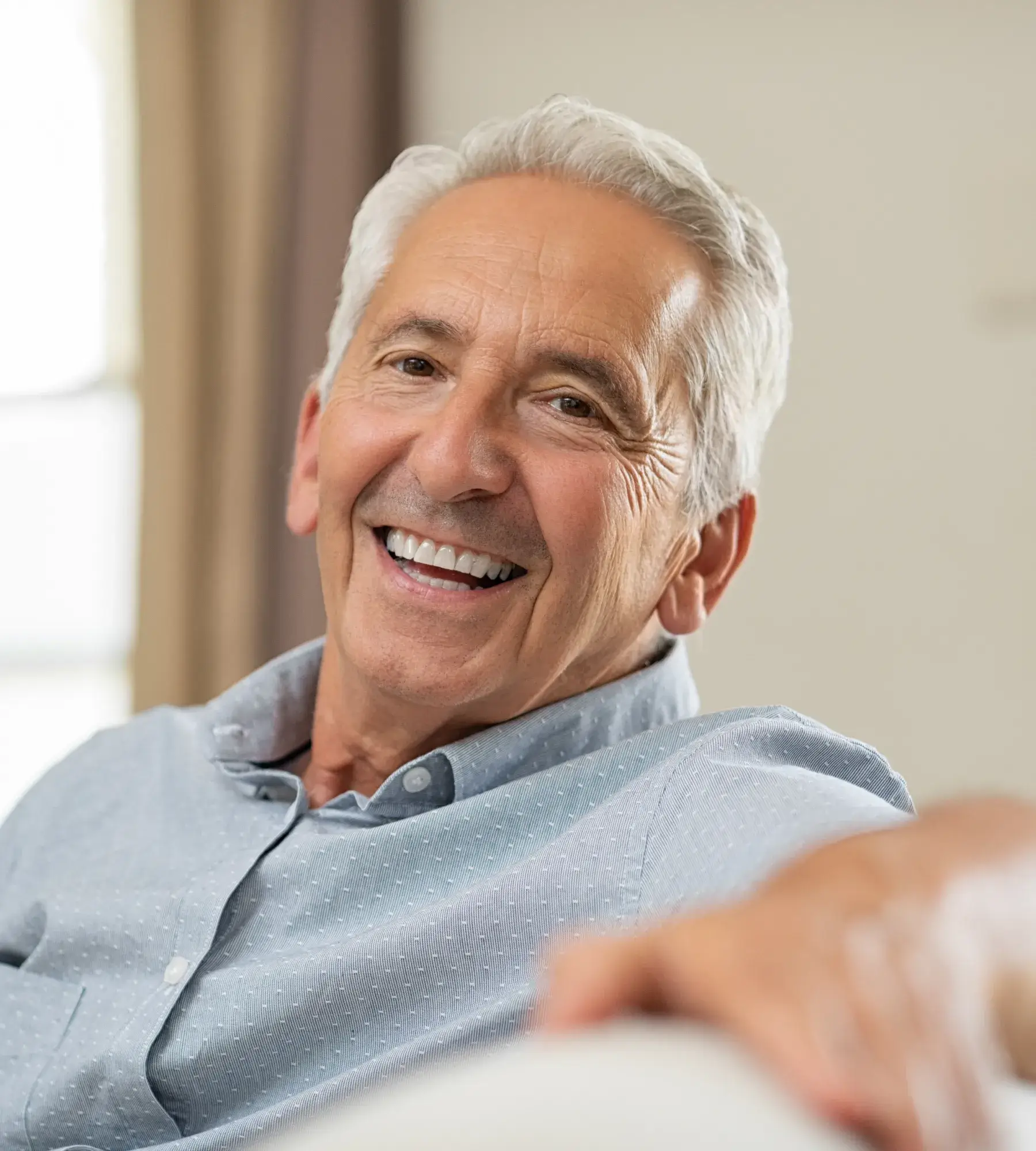 Smiling elderly man with gray hair wearing a light blue shirt sitting comfortably indoors.