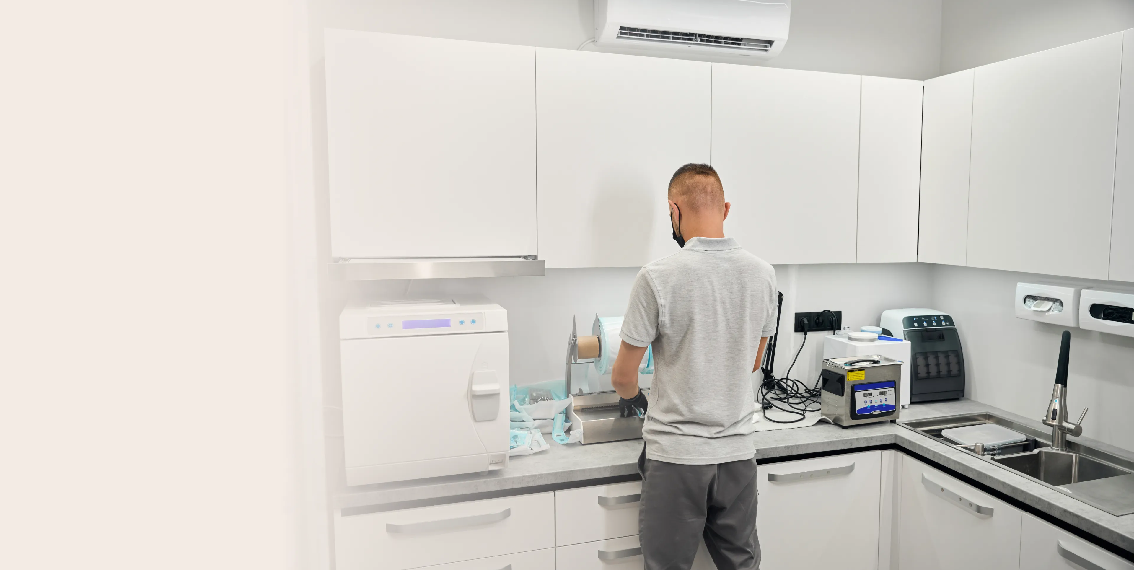 Person wearing a mask and gloves working in a modern sterilization room with medical equipment and white cabinets.