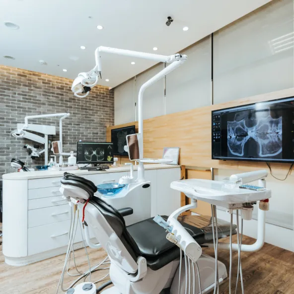Modern dental clinic room with reclining dental chair, overhead light, dental tools, and screens displaying dental X-rays.