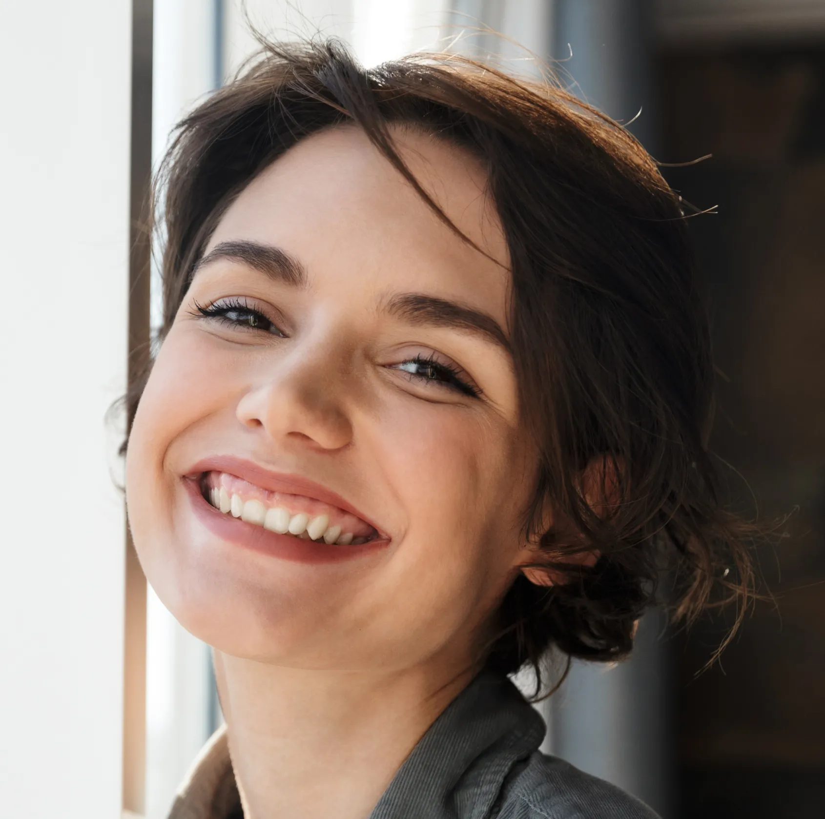 Close-up portrait of a young woman with short dark hair smiling warmly by a window.