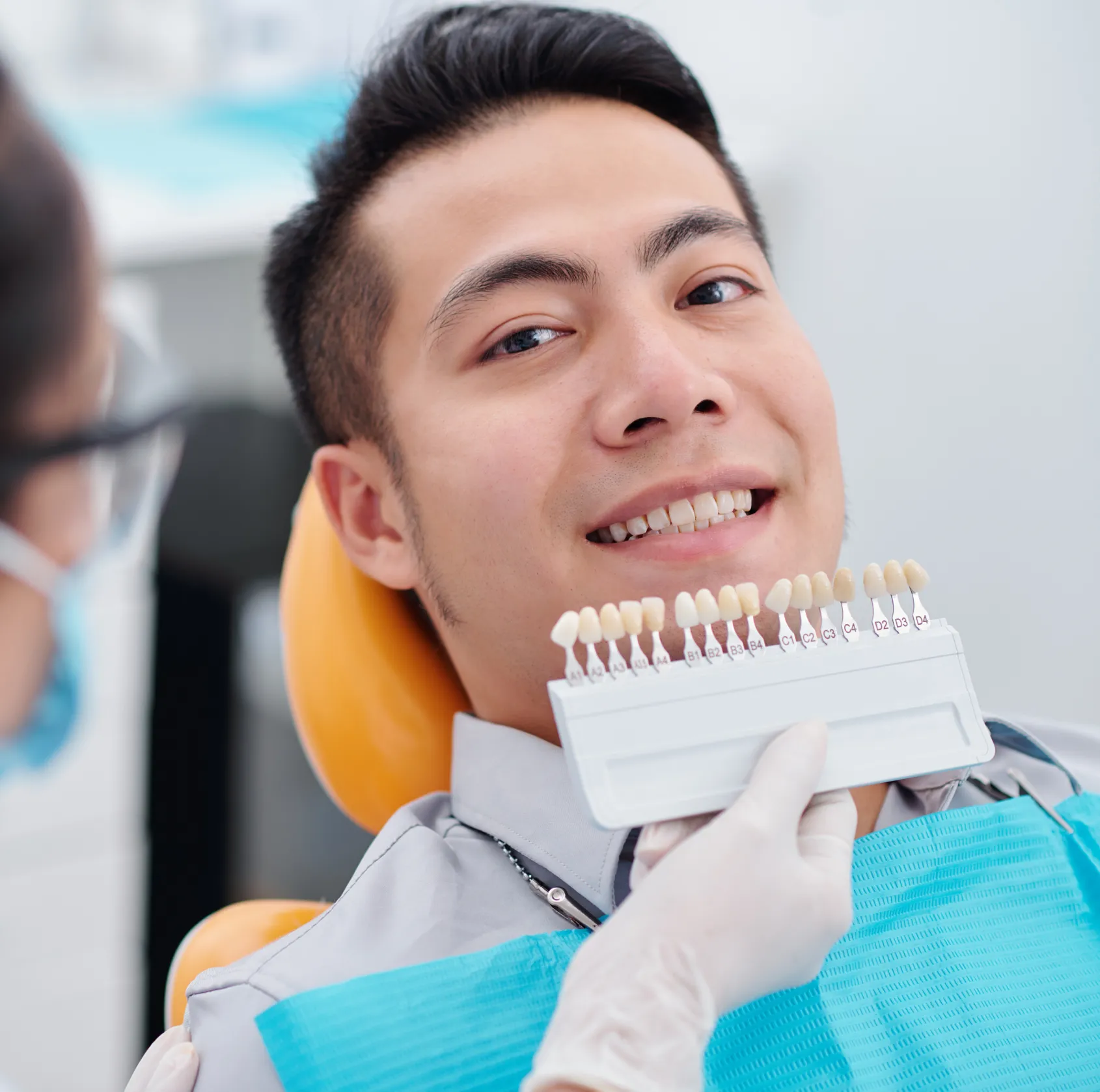 Dentist holding a tooth shade guide next to a smiling man's teeth in a dental clinic.