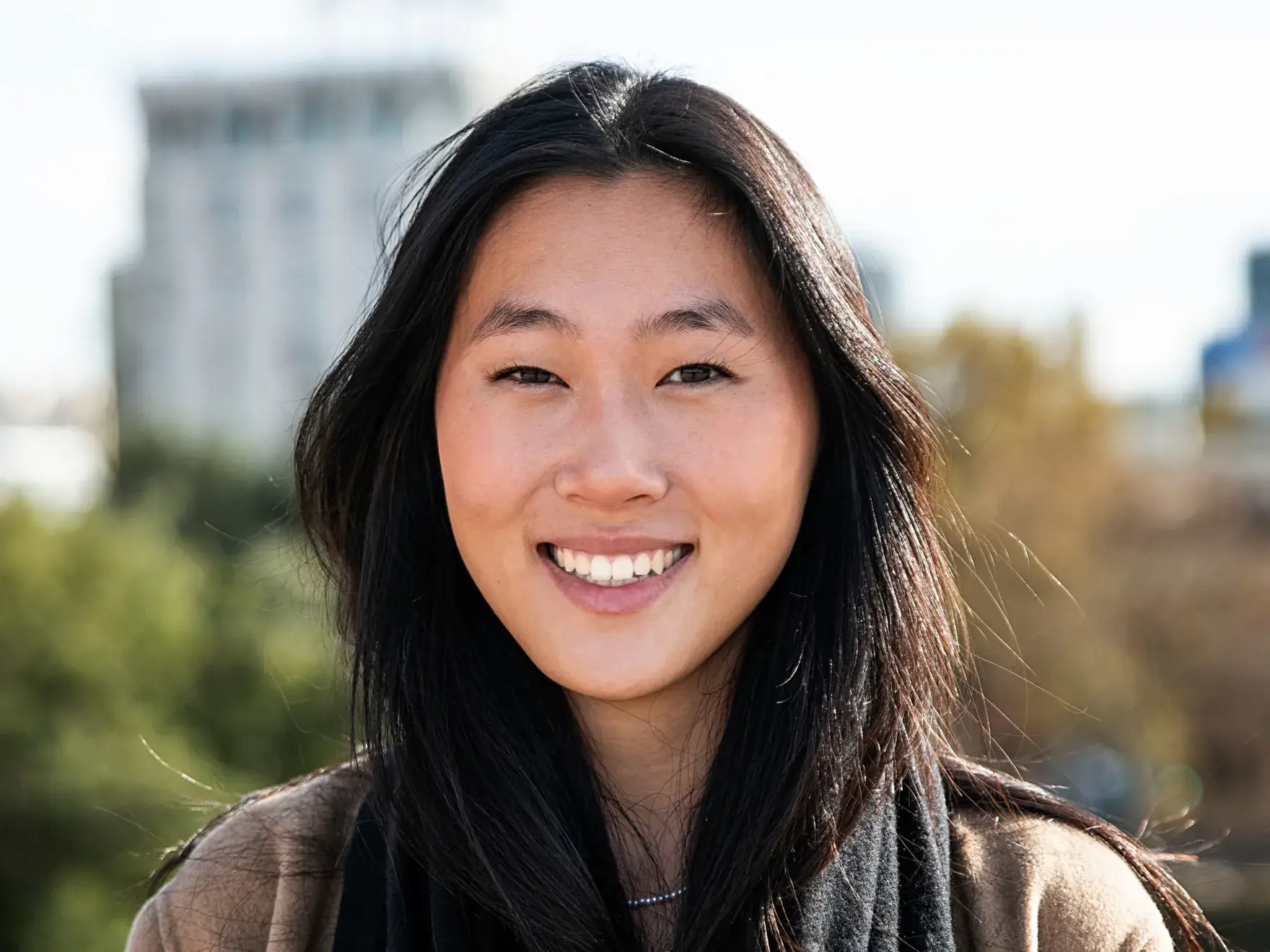 Smiling woman with long black hair standing outdoors with a blurred cityscape background.