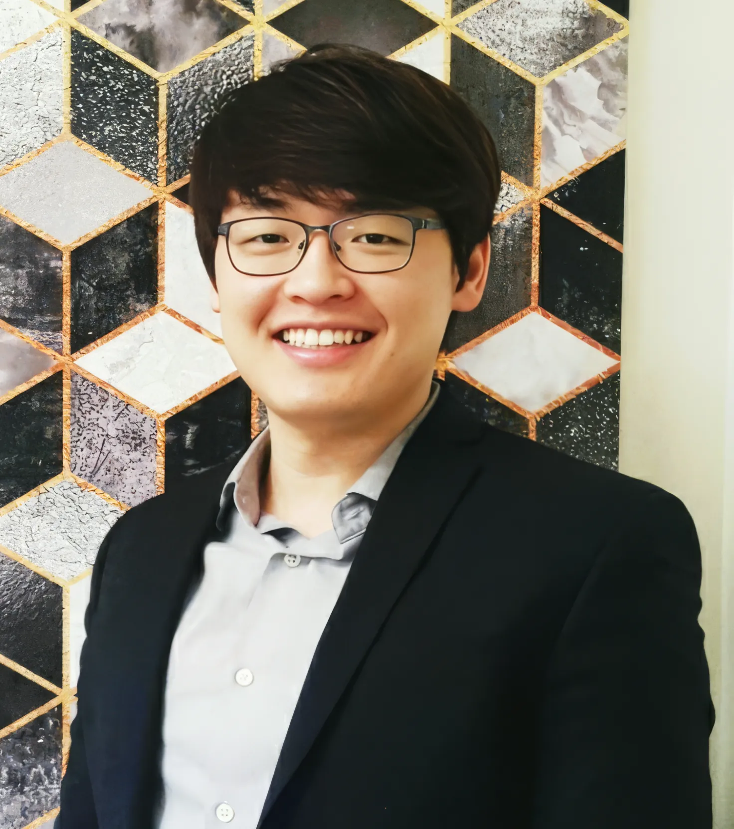 Smiling young man with glasses wearing a dark blazer and light shirt standing in front of a geometric patterned wall.