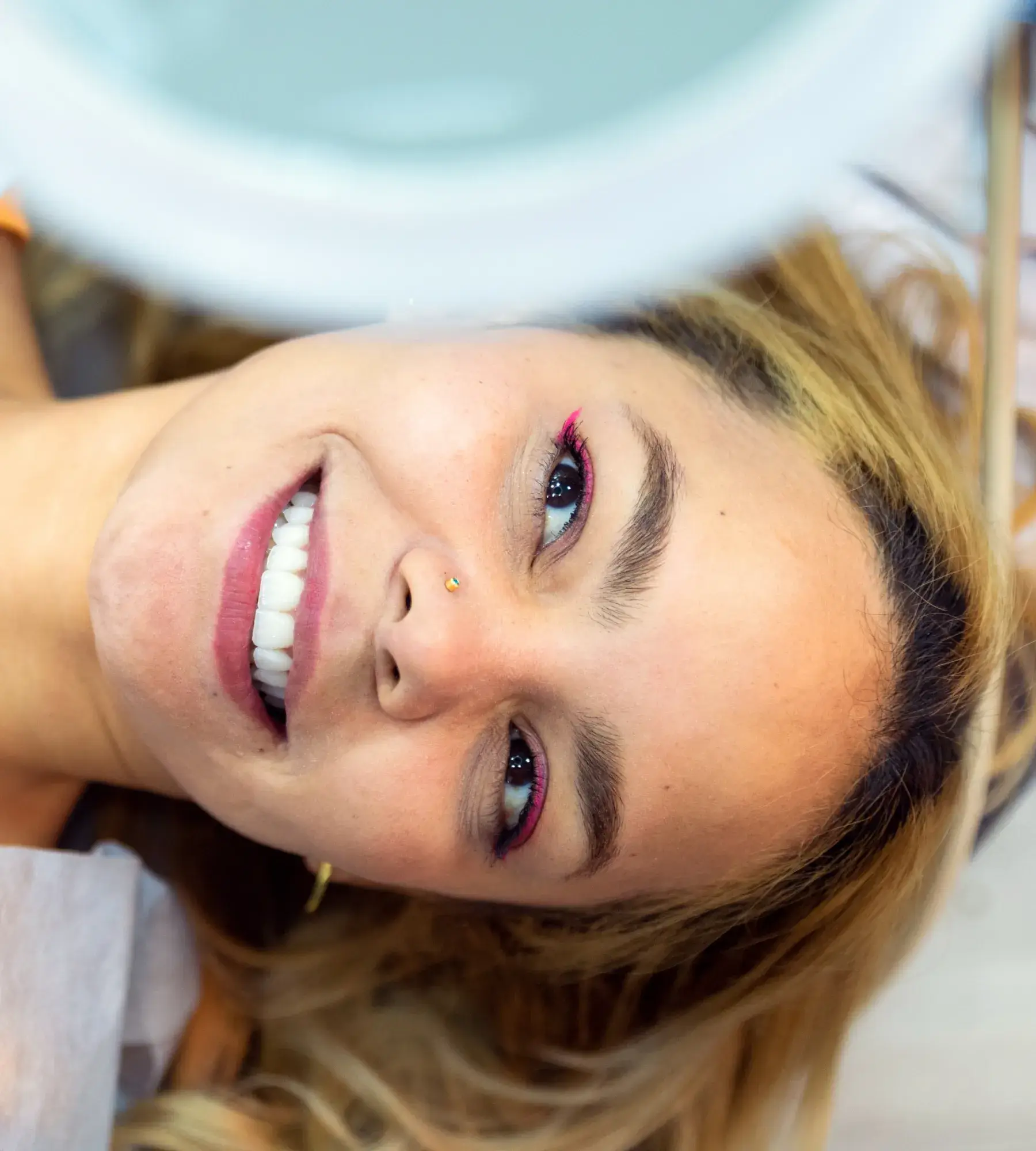 Close-up of a smiling woman with blonde hair, pink eyeliner, and a nose stud lying down with a blurred light above her.