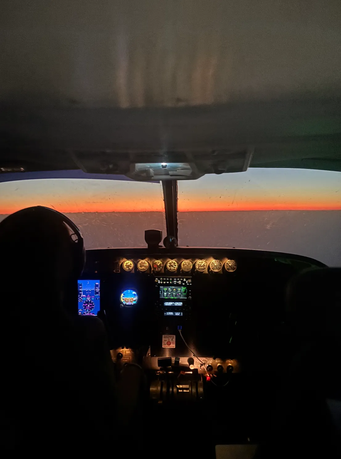 Cockpit interior of a small airplane at dusk with illuminated instruments and a pilot wearing headphones.