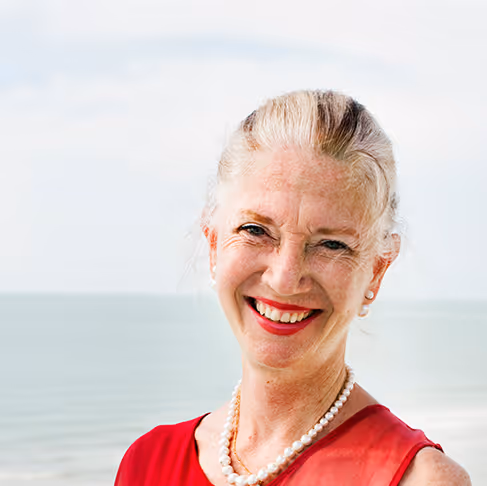 Smiling older woman with light hair wearing a red sleeveless top and pearl necklace, standing outdoors near the sea.