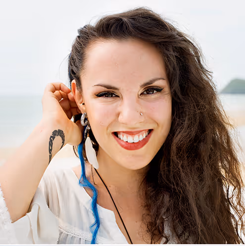 Smiling woman with long curly hair, a nose ring, a feather earring, and a visible tattoo on her wrist.