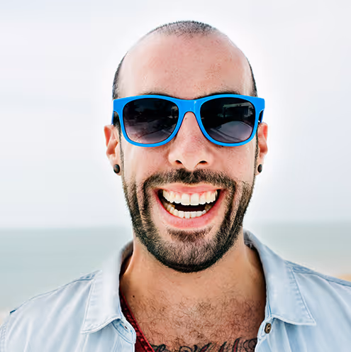 Smiling man with beard and blue sunglasses wearing a light denim shirt at the beach.