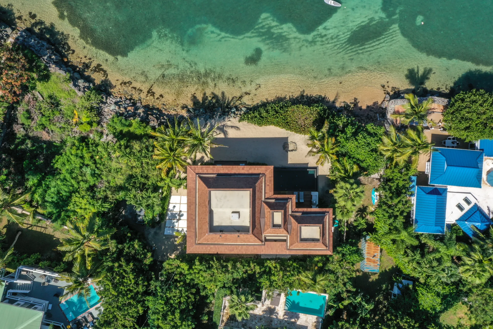 Aerial view of a beachfront house with a terracotta roof surrounded by palm trees and greenery, next to clear turquoise water and a sandy beach.