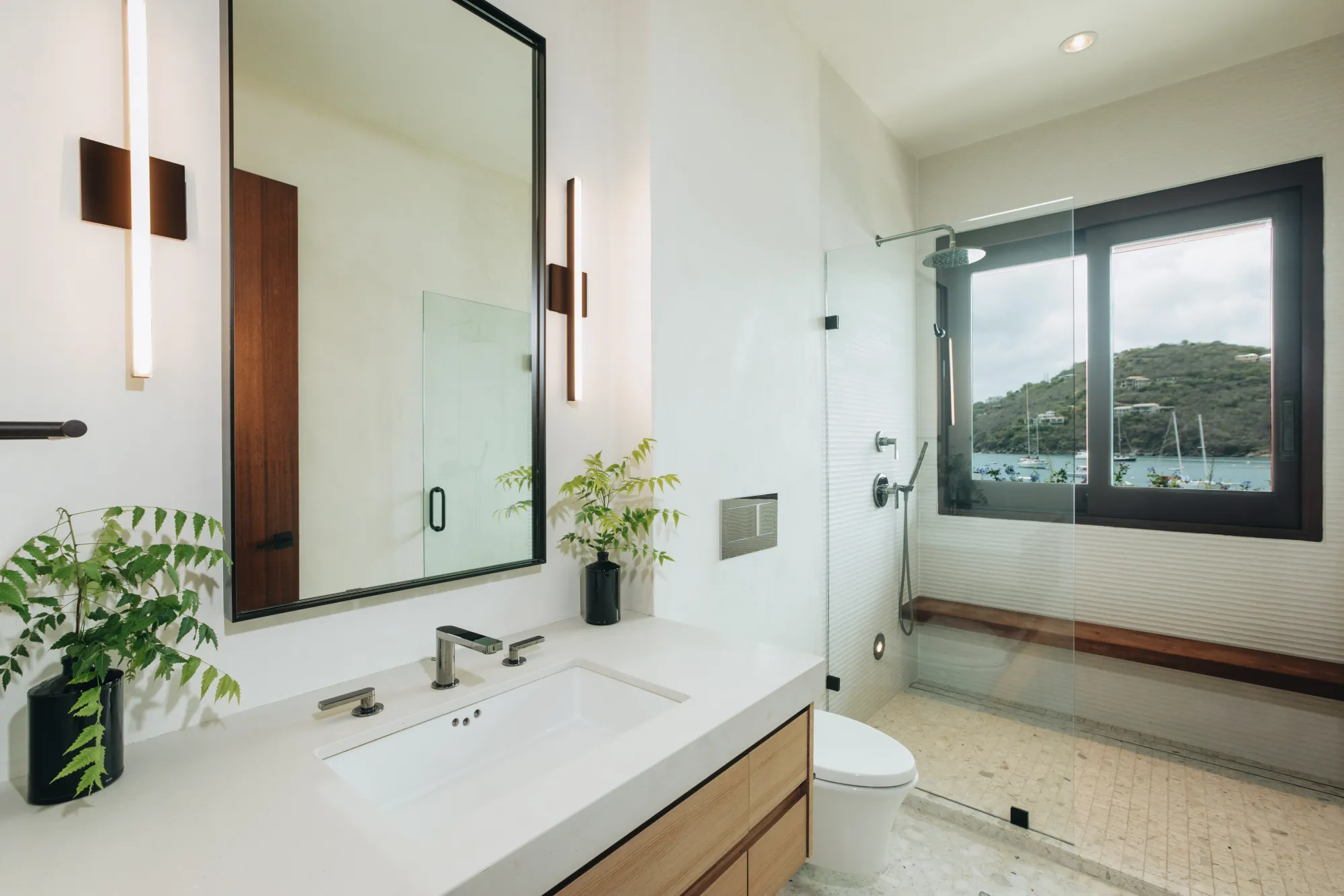 Modern bathroom with white sink, wooden cabinets, glass-enclosed shower, and large window showing boats and hills outside.