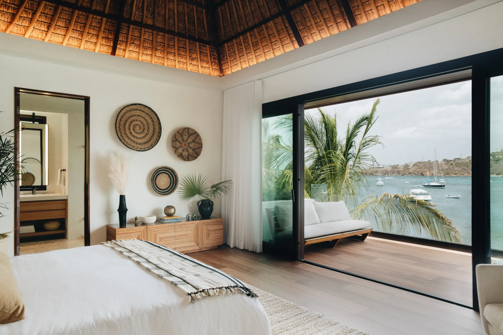 Modern bedroom with white bed, wooden furniture, decorative wall baskets, and sliding glass doors opening to a balcony with seating and ocean view.