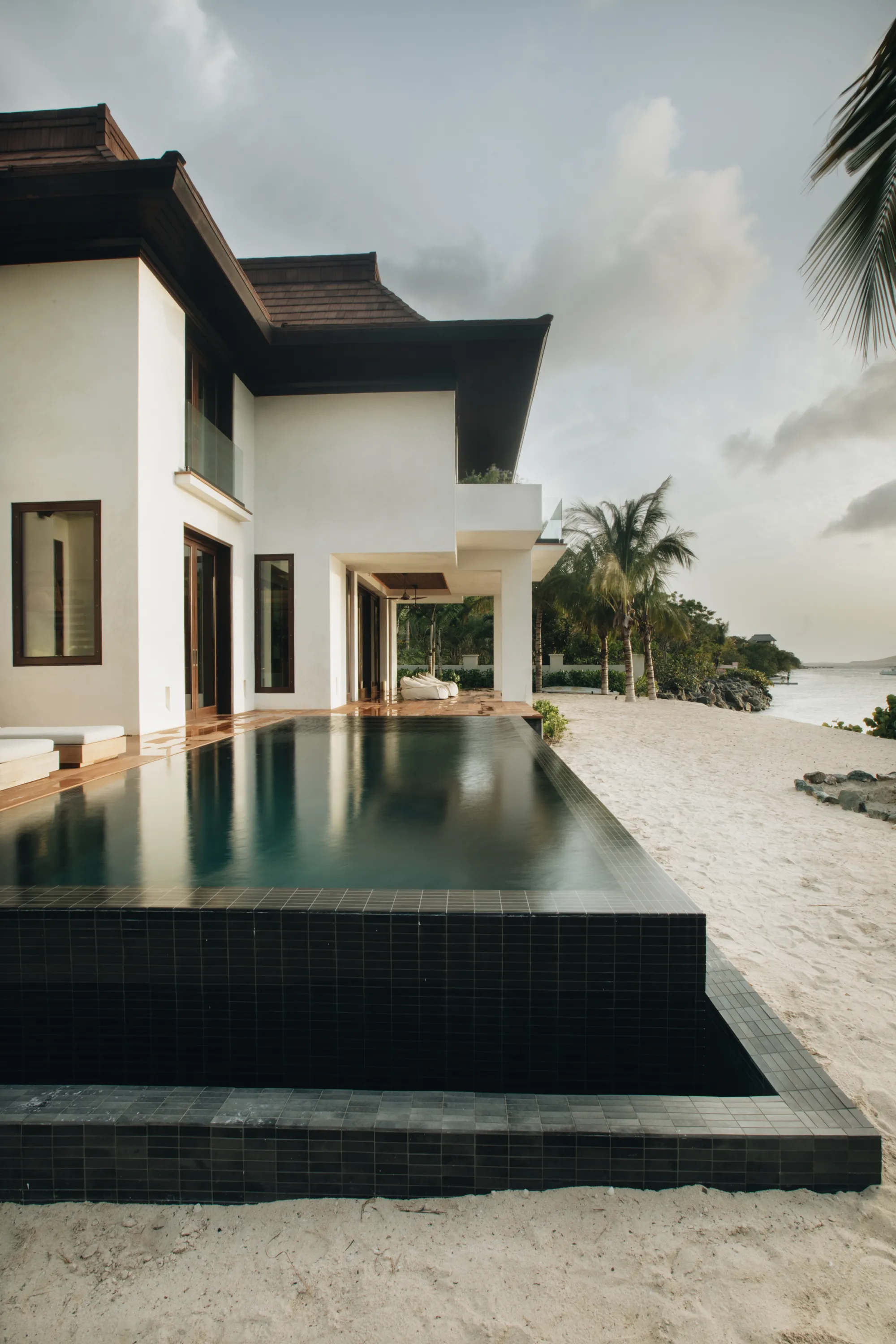 Modern white beachfront house with an infinity pool overlooking sandy beach and palm trees.