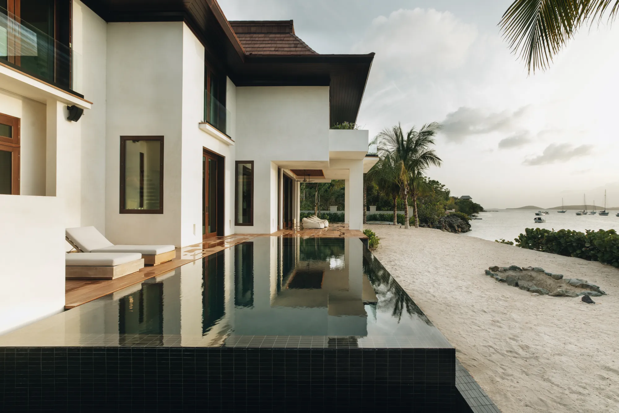 Modern white house with infinity pool next to a sandy beach and palm trees overlooking a calm bay with boats.