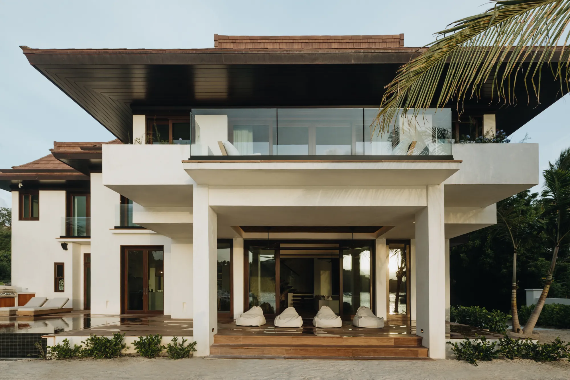 Modern two-story white house with large glass doors, balcony, and outdoor seating on a sandy ground.
