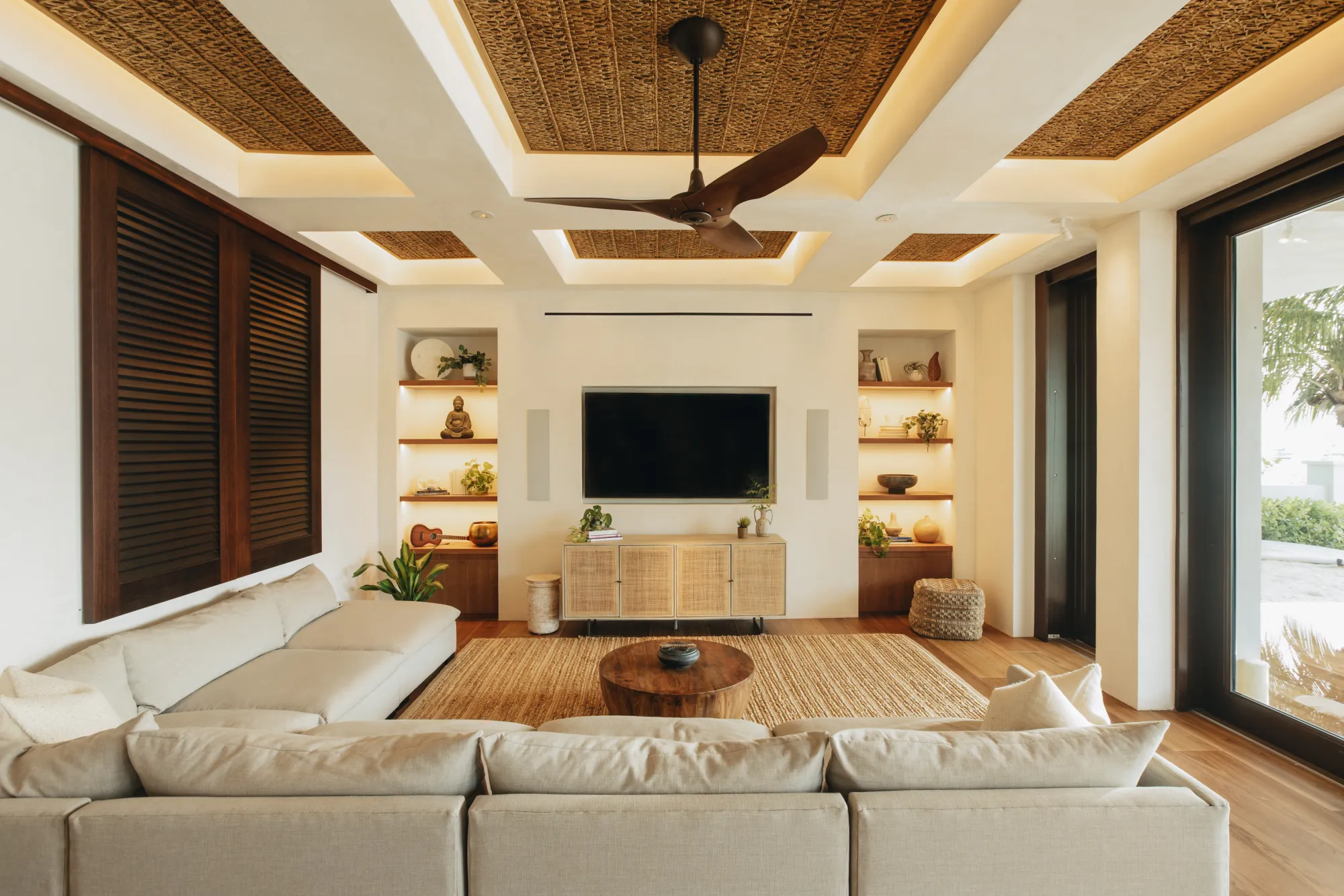 Modern living room with beige sofas, wooden coffee table, woven rug, ceiling fan, built-in shelves with decor, and large glass doors.