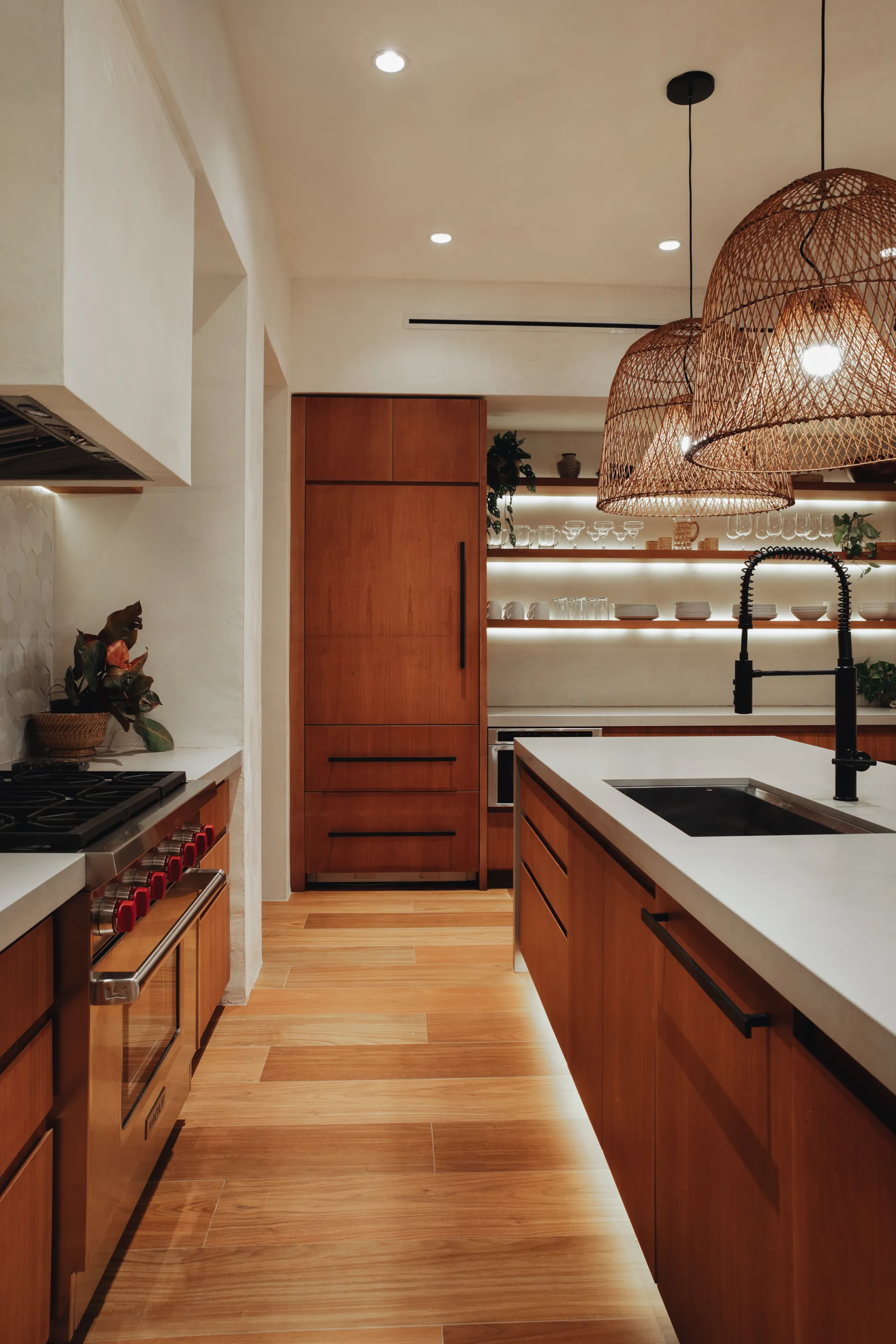 Modern kitchen with wooden cabinets, white countertops, black faucet, hanging wicker pendant lights, and open shelves with glassware and plates.