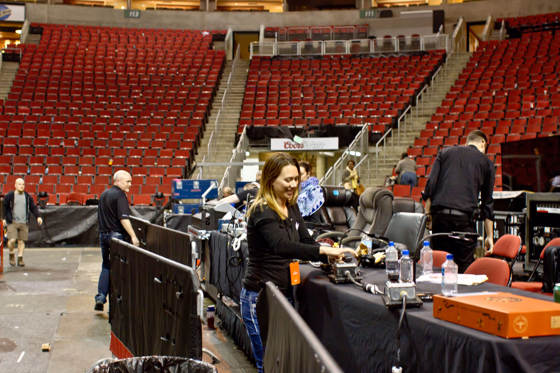 People working behind a black-covered table with chairs and equipment in an empty arena with red seats.