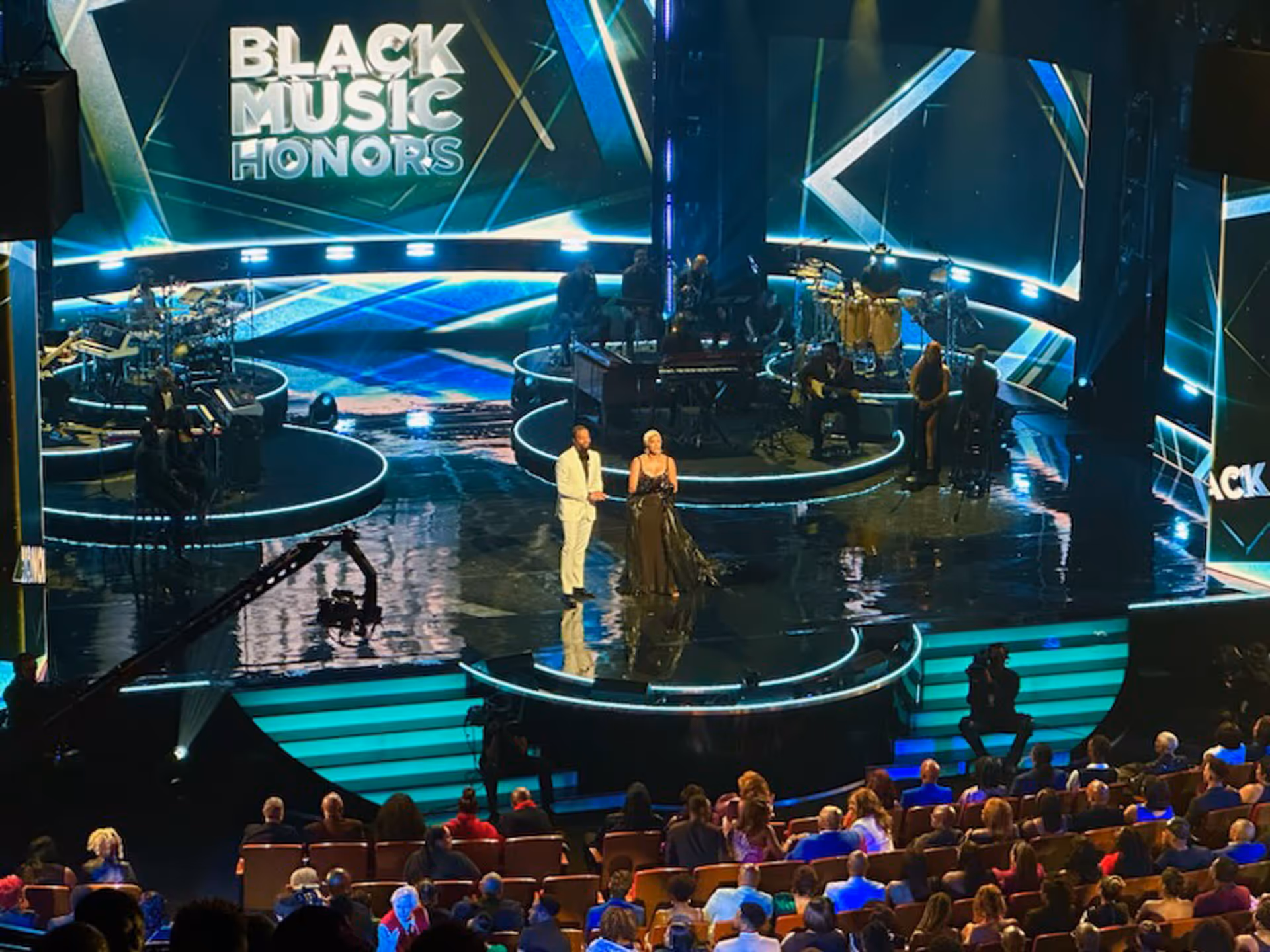 Hosts standing on stage with musicians at the Black Music Honors event with an audience watching.