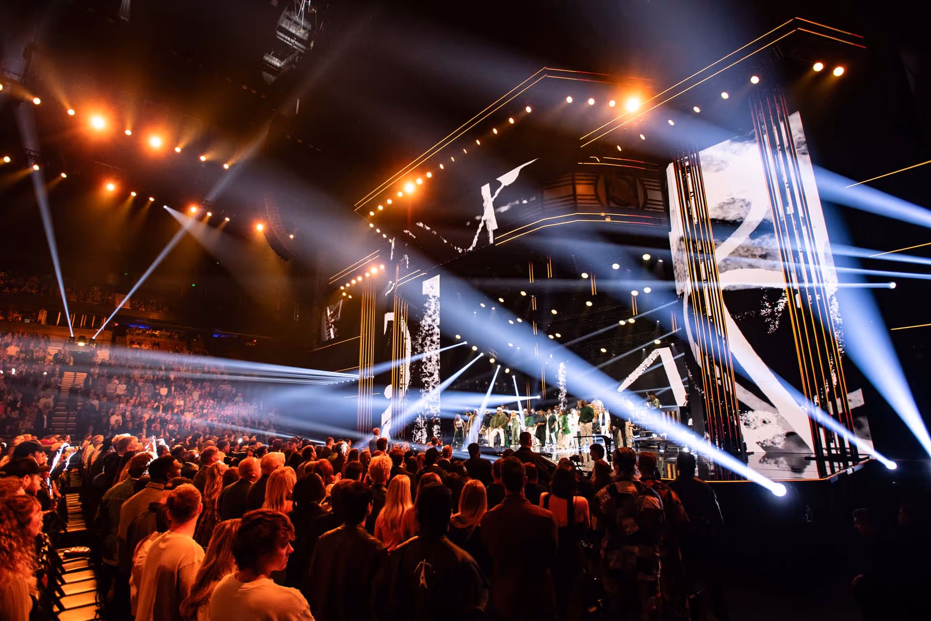 Large crowd watching a brightly lit stage with performers and multiple vertical LED screens in an indoor concert venue.