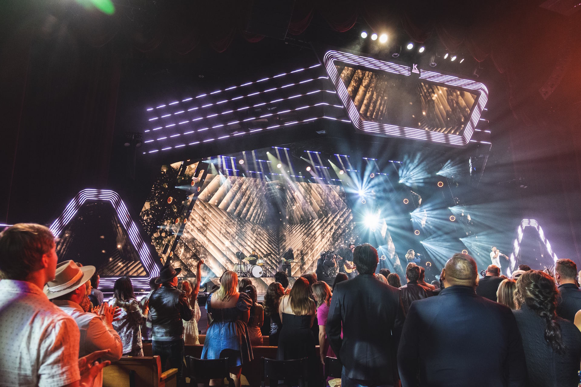 Outdoor concert with a large crowd watching a band performing on a stage with two video screens showing a singer.