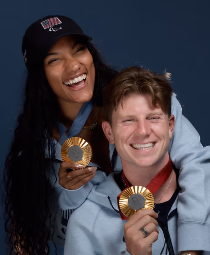 Two smiling athletes holding gold medals, the woman wears a black cap with an American flag and Paralympic symbol.