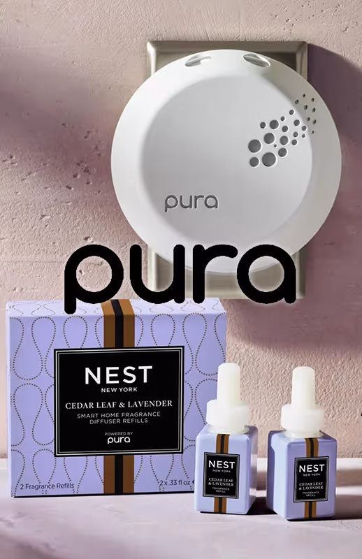White Pura smart home fragrance diffuser plugged into a wall outlet with two Nest New York cedar leaf and lavender fragrance refill bottles and packaging placed below it.
