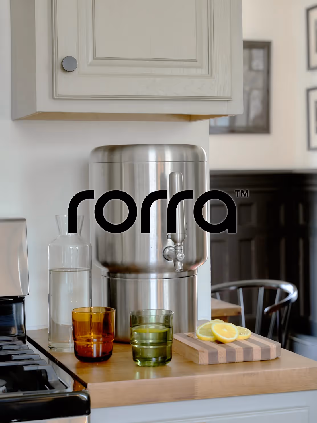 Stainless steel water dispenser on kitchen counter with glass carafe, colored drinking glasses, and sliced lemons on a cutting board.
