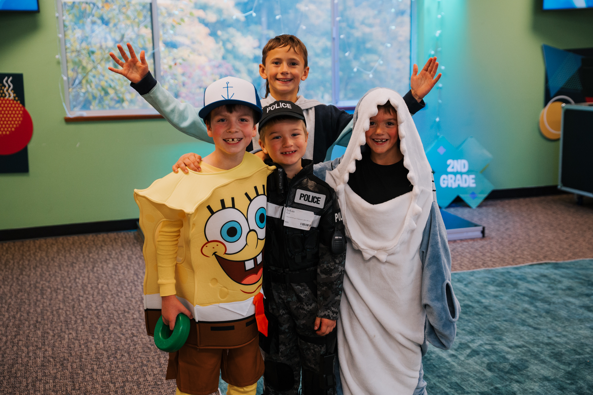 Four smiling children dressed in costumes, and a boy with a cap raising his hands, standing indoors near a window with a sign saying '2nd Grade'.