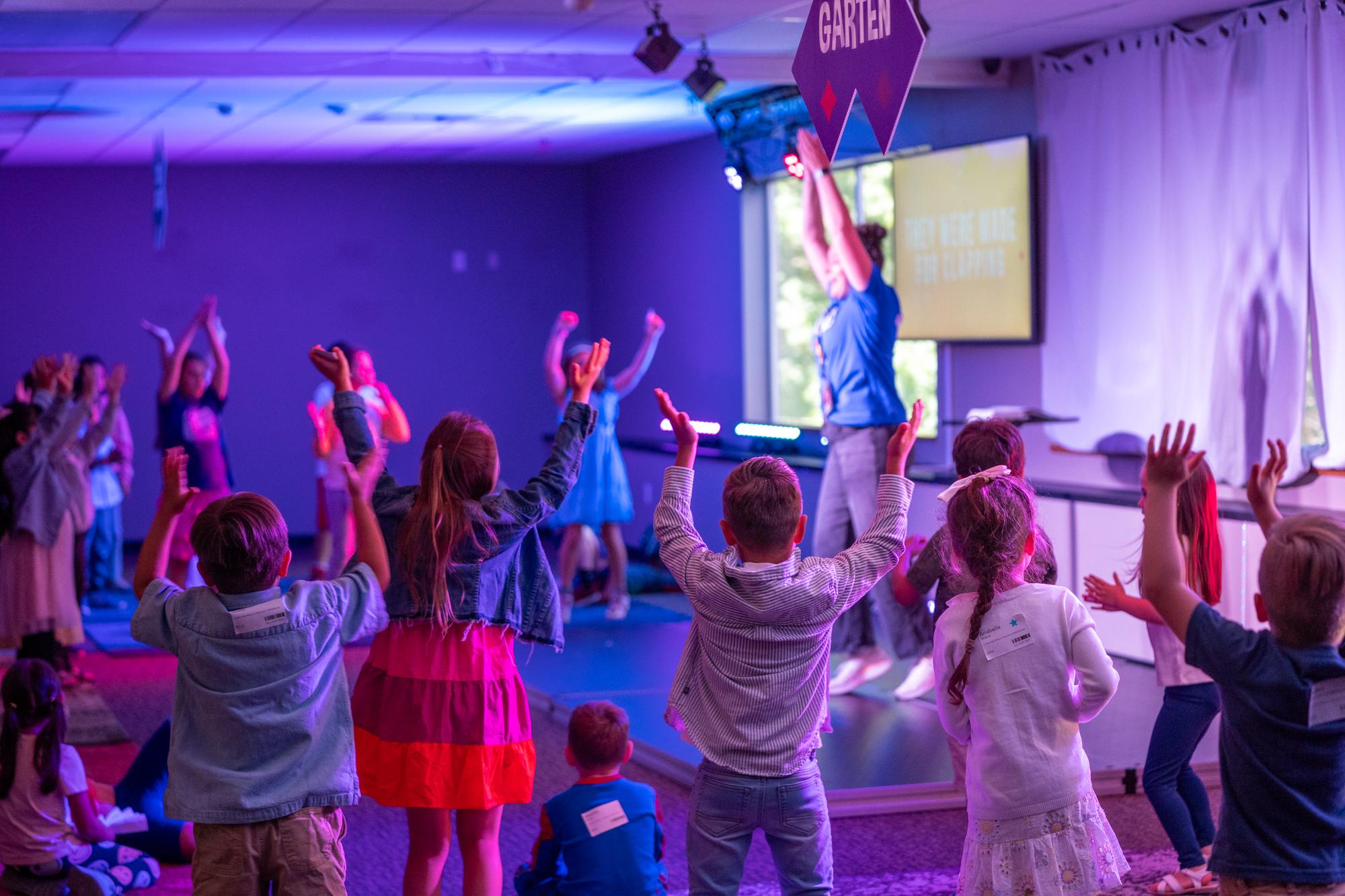 Group of children with raised hands participating in an indoor activity led by an instructor on stage under colorful lights.