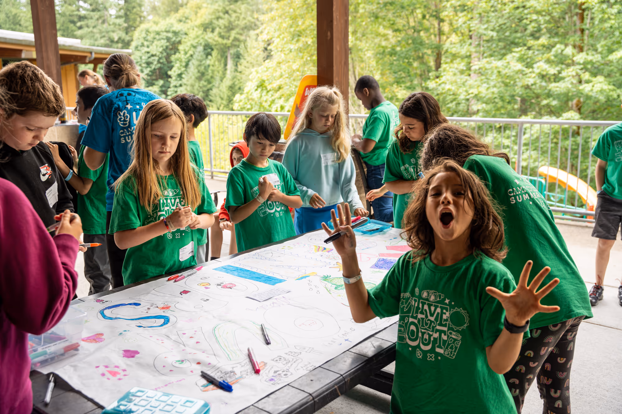 Children wearing green camp t-shirts draw and color on a large table outdoors under a covered area.