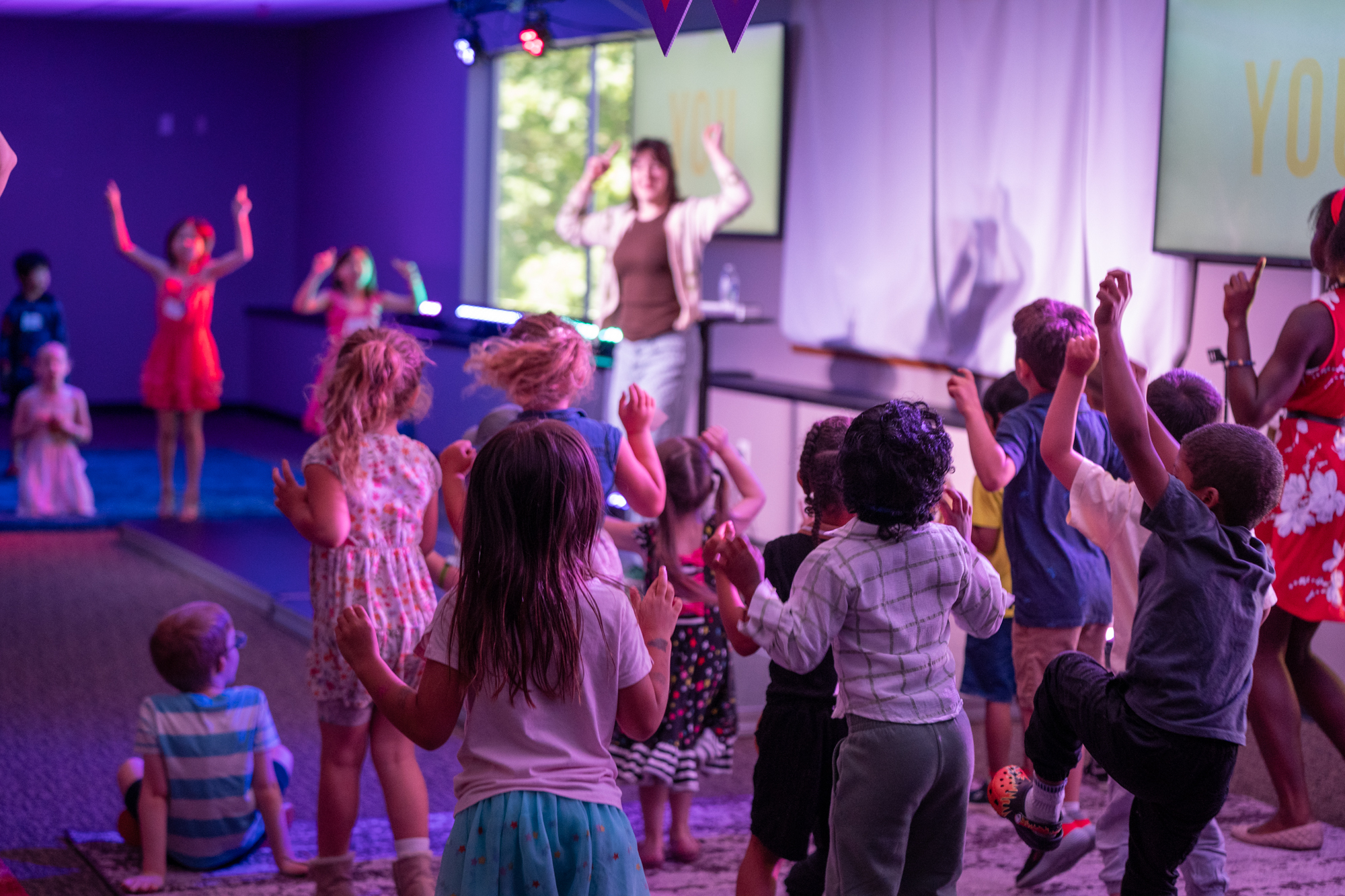 A group of kids gathered around in a classroom in excitement with hands raised.