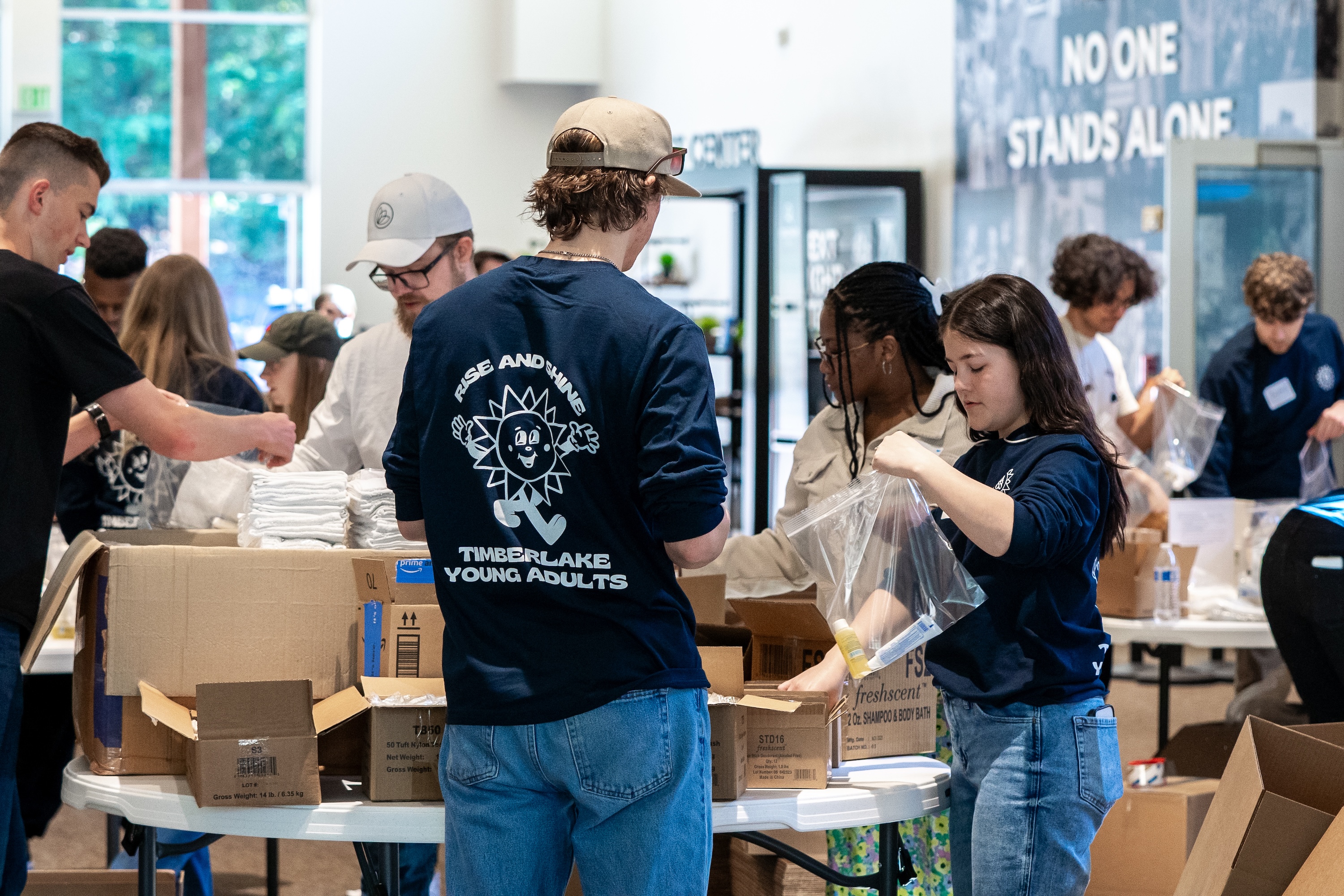 Groups of young adults standing around tables, helping pack materials together.