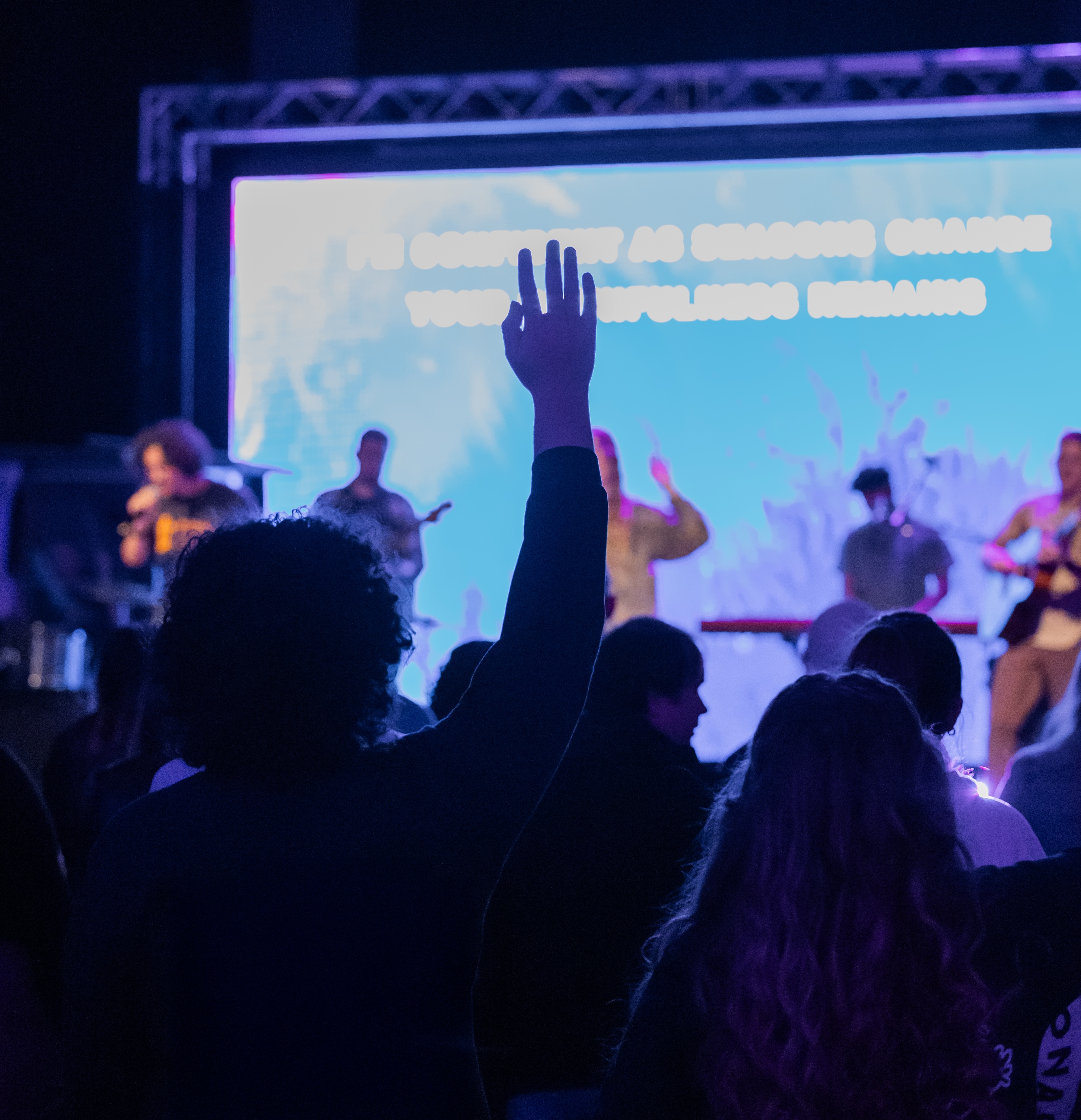 People's arms raised in worship in the foreground with the worship team on stage in the background.