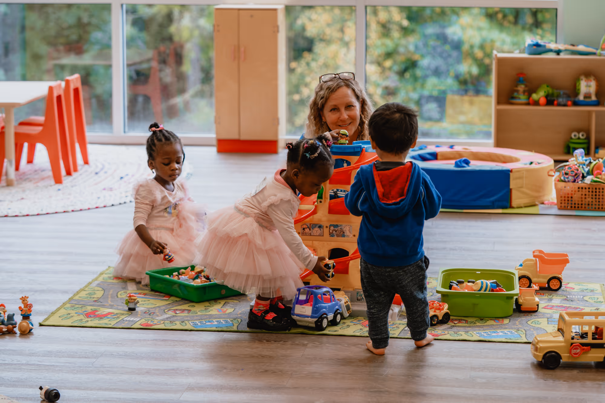 Two young girls in pink tutus and a boy in a blue hoodie playing with toy cars and figures on a colorful play mat, with an adult woman smiling at them indoors.
