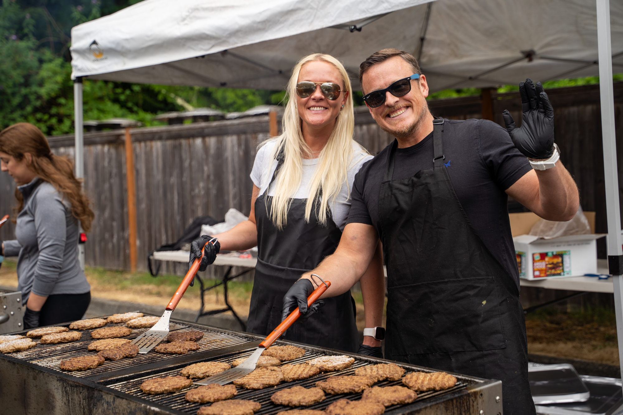 A couple flipping burgers on the grill and smiling at the camera.