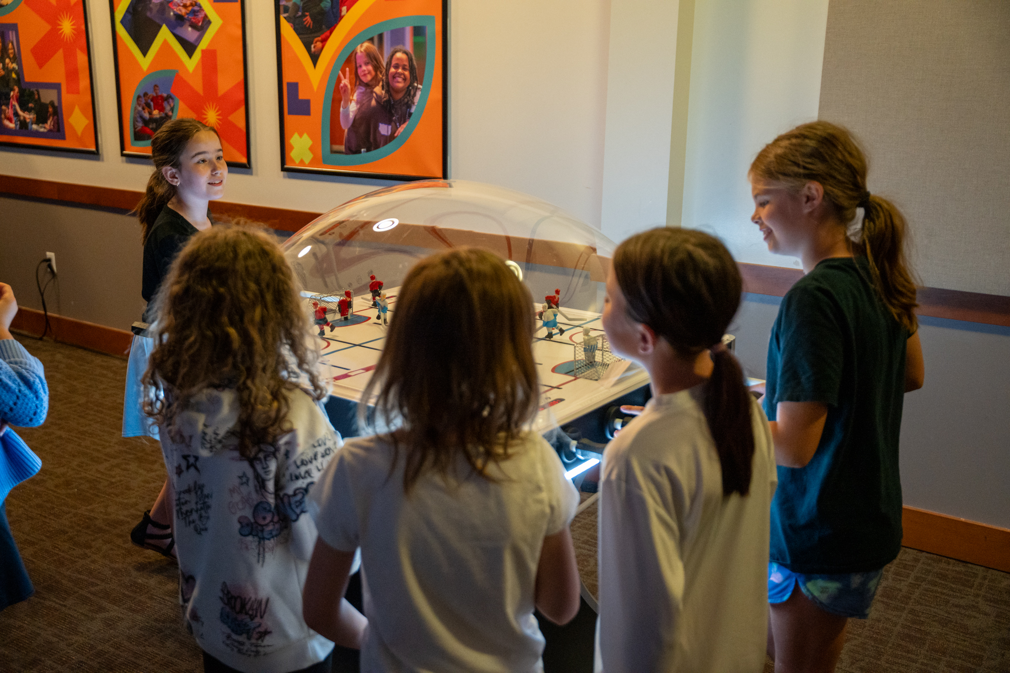 Five children gathered around a hockey game table, playing and watching in a room with colorful photo wall art.