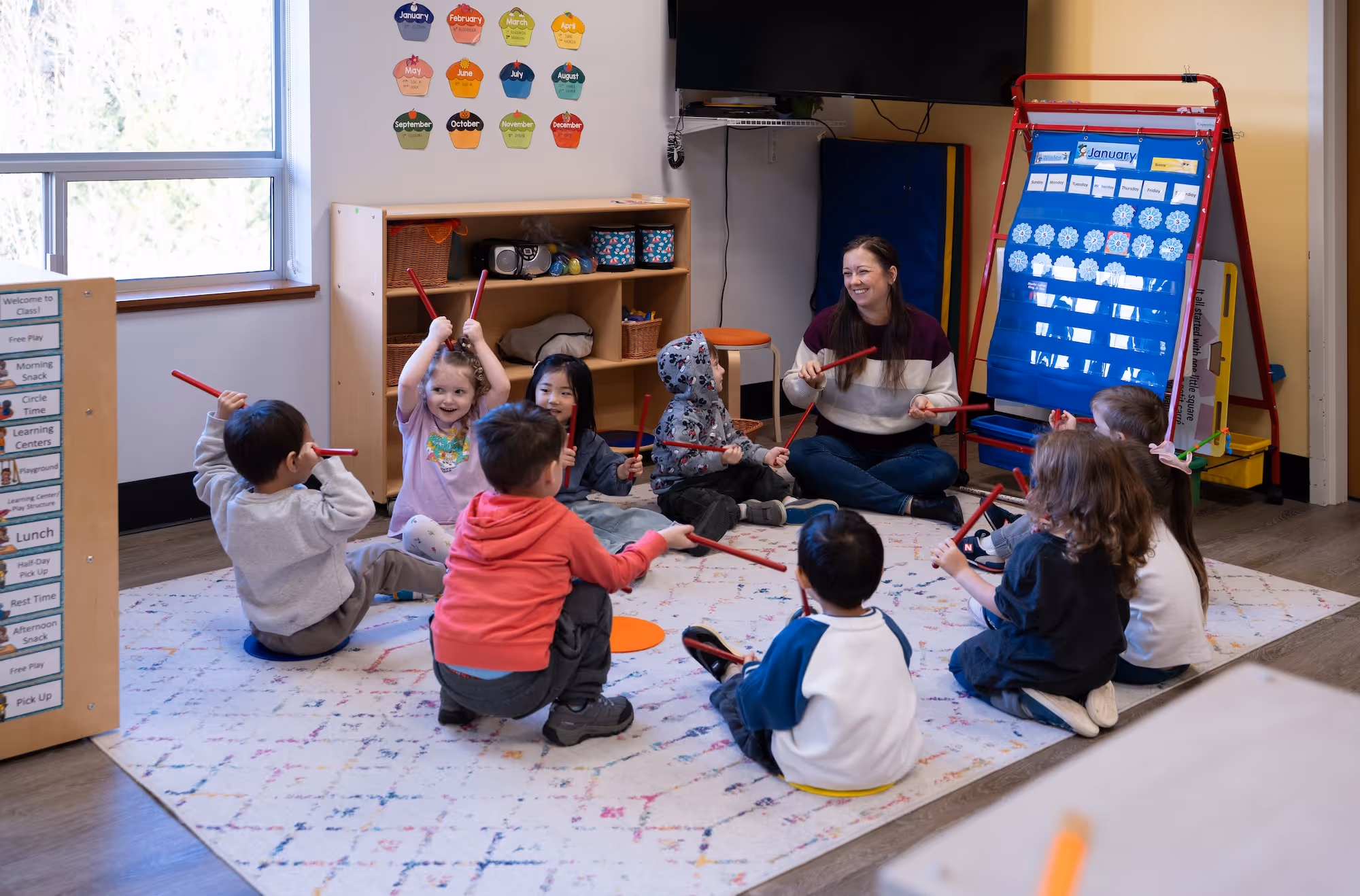 A group of preschoolers sitting on the rug around the teacher, playing with sticks.