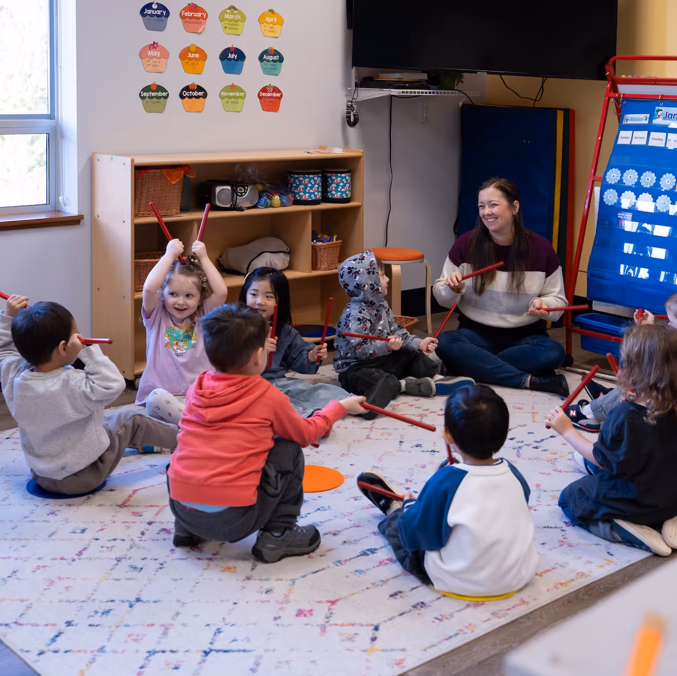 A group of preschoolers sitting on the rug around the teacher, playing with sticks.