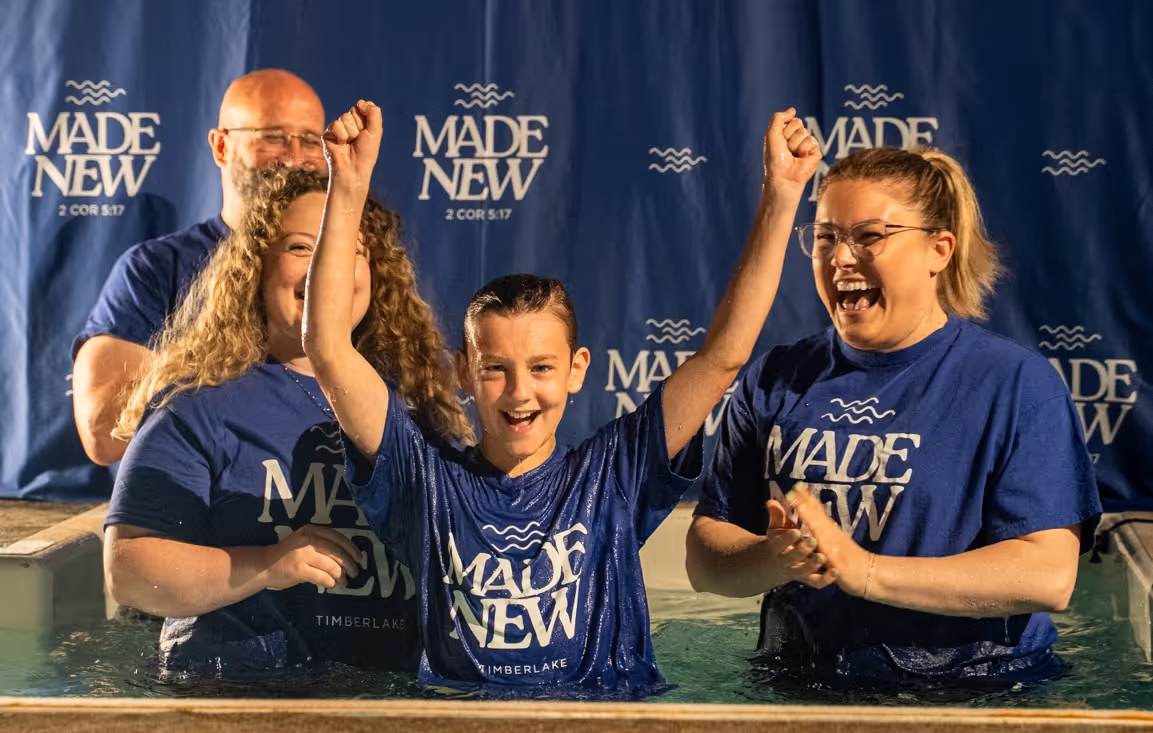Smiling boy with raised arms celebrating baptism in water, surrounded by three adults wearing blue 'Made New' shirts.