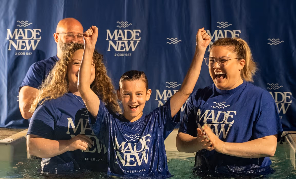 Smiling boy with raised arms celebrating baptism in water, surrounded by three adults wearing blue 'Made New' shirts.