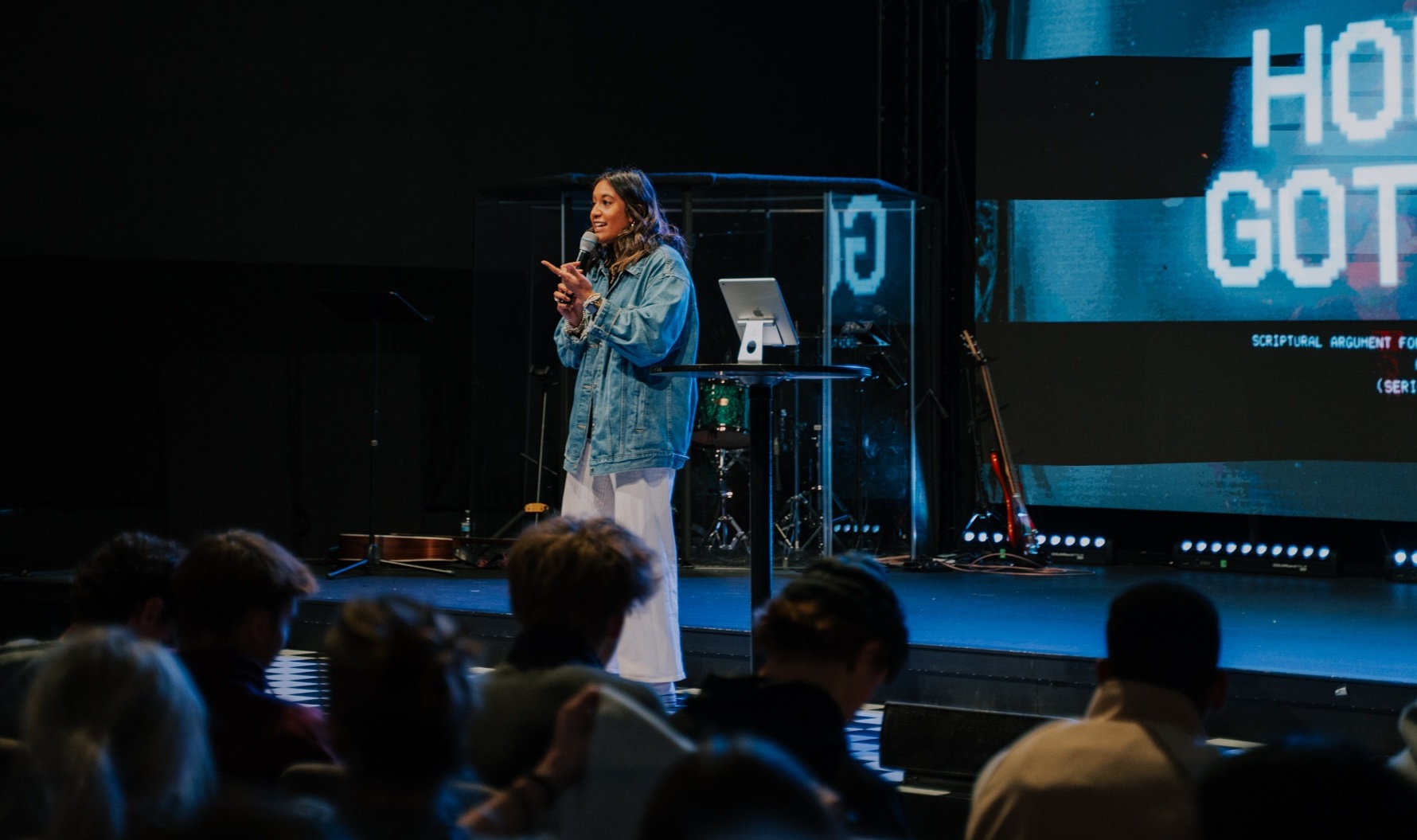 A youth pastor preaching on stage with a large audience of highschoolers listening intently.