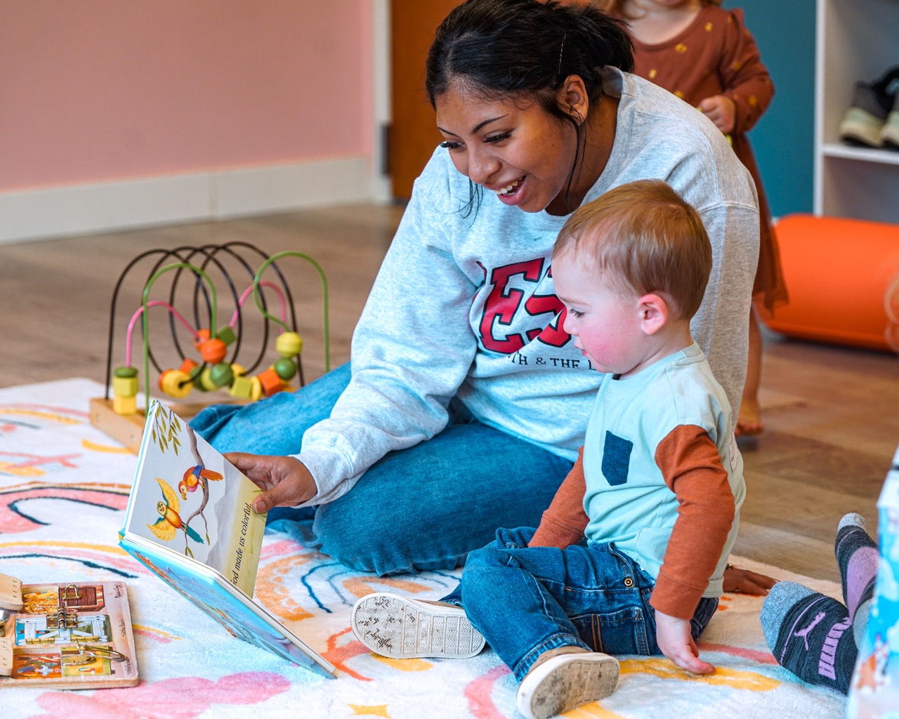 Woman reading a colorful picture book to a toddler seated on a patterned rug, with toys in the background.