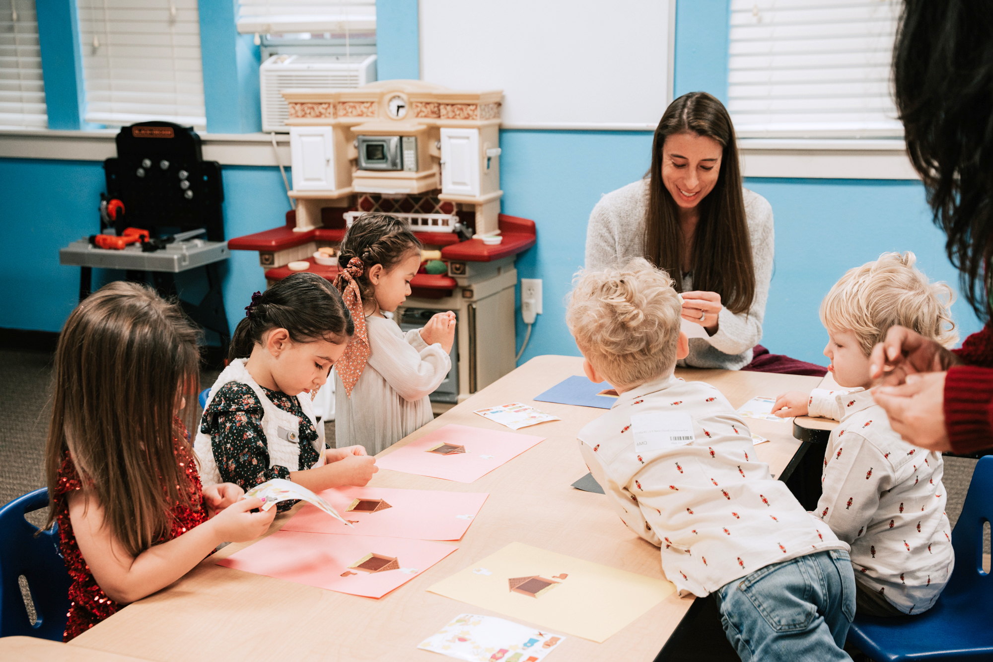 A group of young children sitting around a table doing arts and crafts with two adult women assisting.